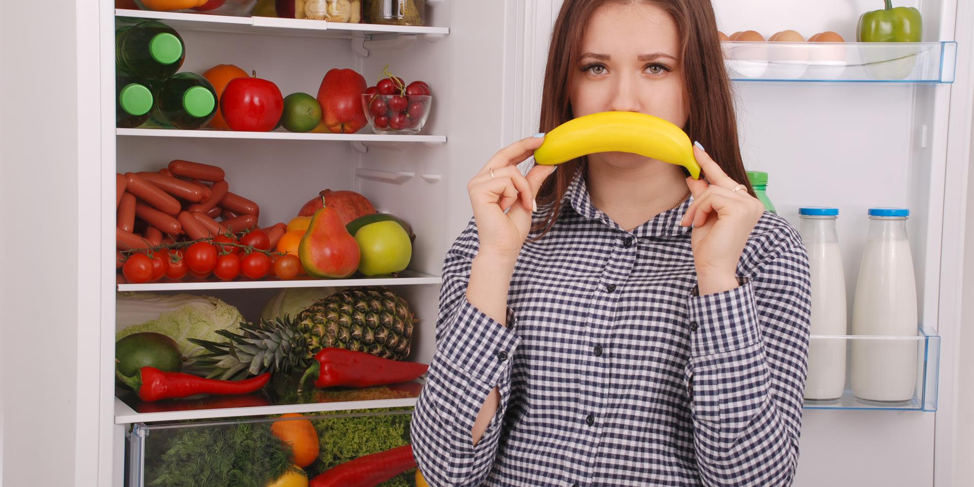 בחורה עומדת מול מקרר מלא פירות וירקות ומחזיקה בננה בצורת חיוך הפוך